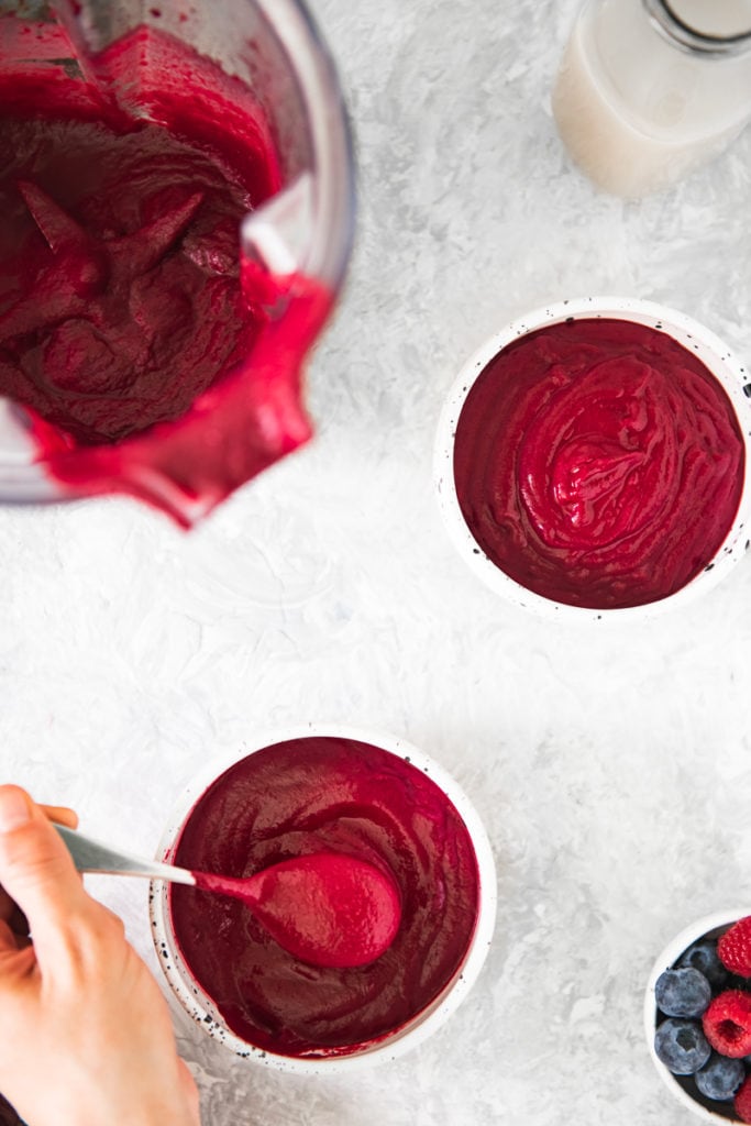 A spoonful of a smooth beetroot smoothie bowl being scooped out of a bowl beside another full bowl, an almost empty blender and a little bowl of berries