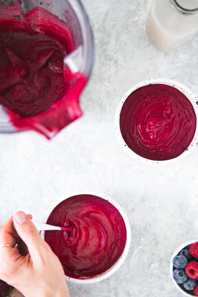 Two beet smoothie bowls with a hand dipping a spoon in one and the blender and a bowl of mixed berries sitting sitting beside