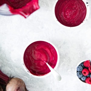 Two beet smoothie bowls with a hand dipping a spoon in one and the blender and a bowl of mixed berries sitting sitting beside