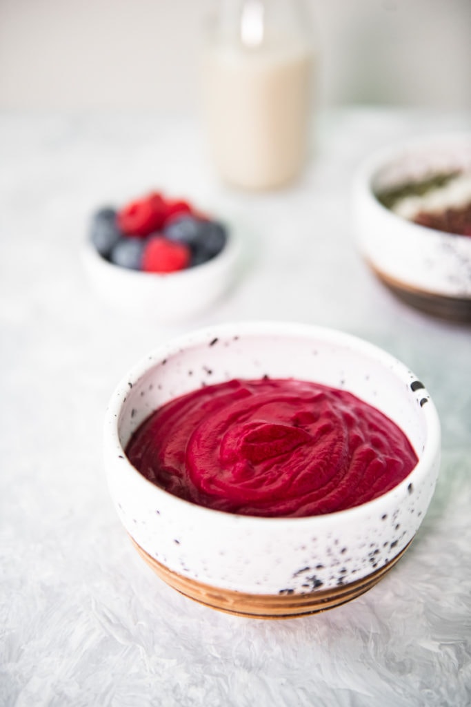 A vibrant berry and beet smoothie bowl front and centre with another smoothie bowl full of toppings, a bowl of mixed berries and bottle of plant-milk behind.