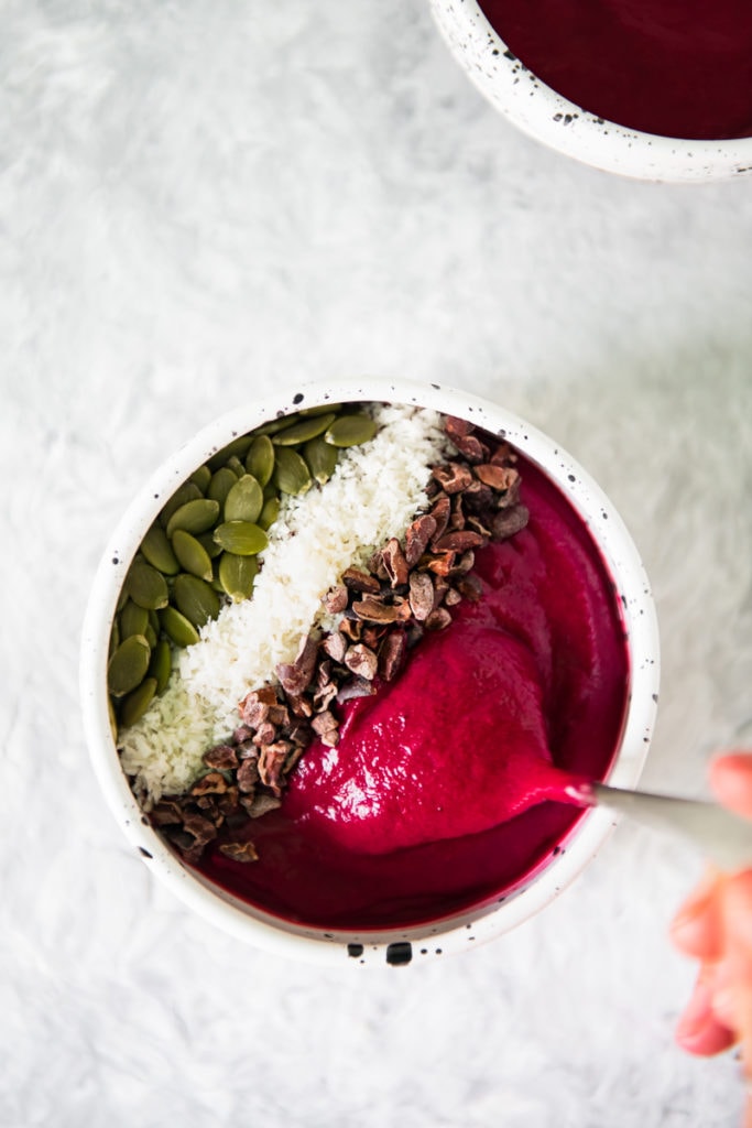 A spoonful of berry beet smoothie bowl being lifted out of a full bowl perfectly topped with cacao nibs, shredded coconut and pumpkin seeds.