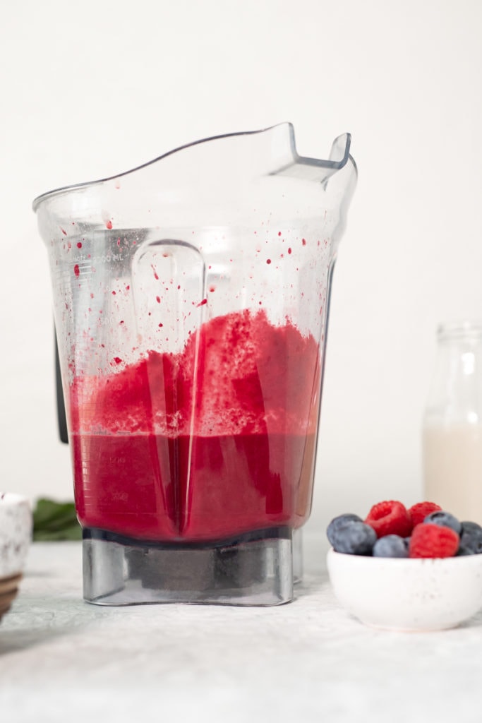 A blender container full of freshly blended cinnamon berry & beetroot smoothie bowl with a bowl of fresh berries and a jar of milk.