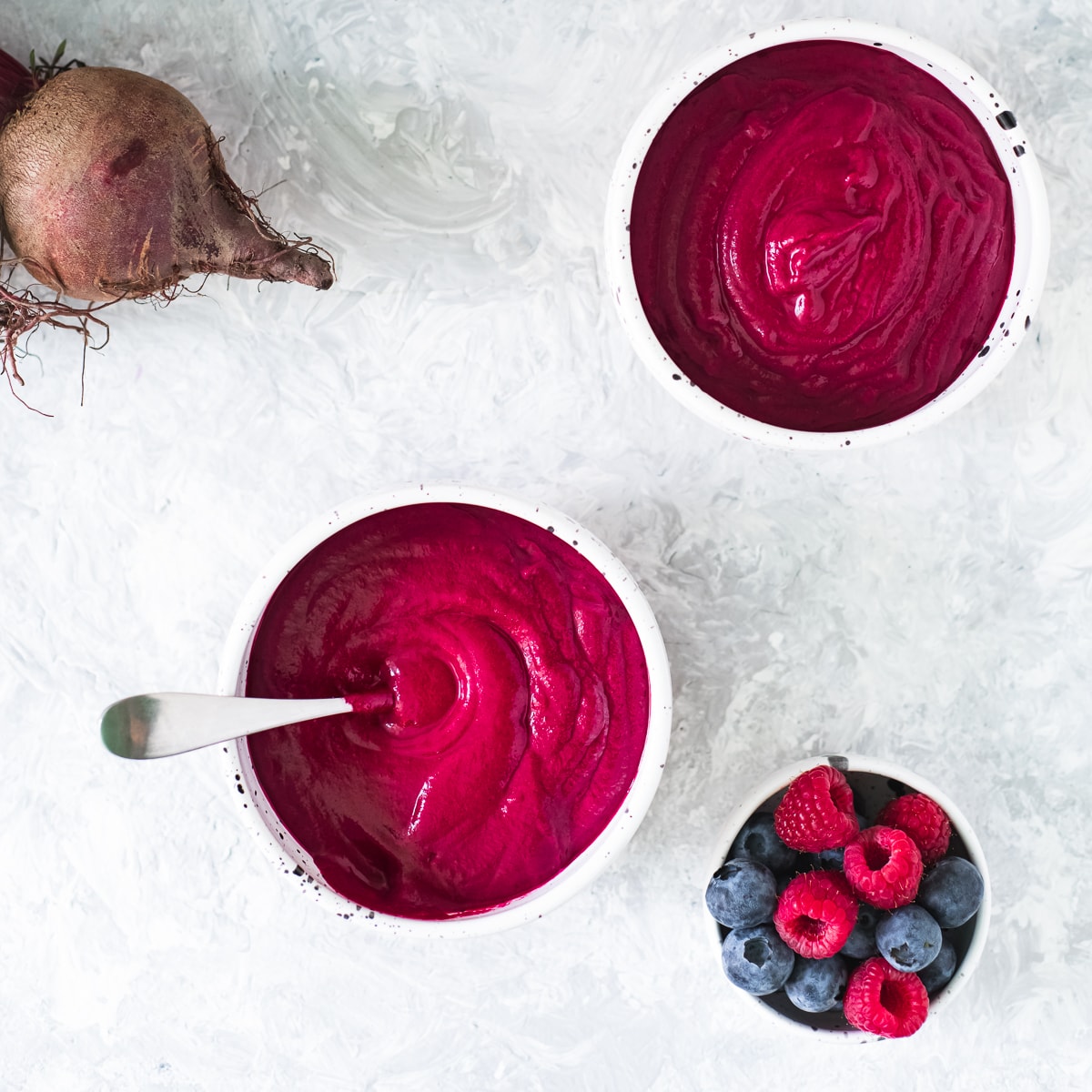 Two deep red beetroot smoothie bowls beside a bowl of berries and a bunch of beets.