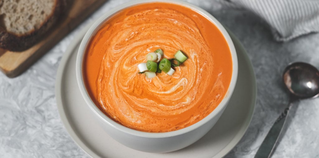 A big bowl of creamy tomato soup swirled with cashew cream and topped with green onions. Beside the bowl is a rustic spoon and a cutting board with whole grain bread.