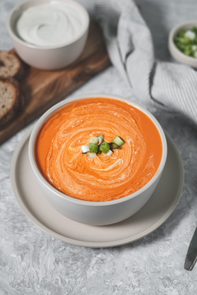 A large bowl of homemade tomato soup swirled with cashew cream and topped with green onion. Surrounding the bowl is a board with cashew cream, bread and on the other side is a linen napkin, spoon and bowl of chopped green onion.