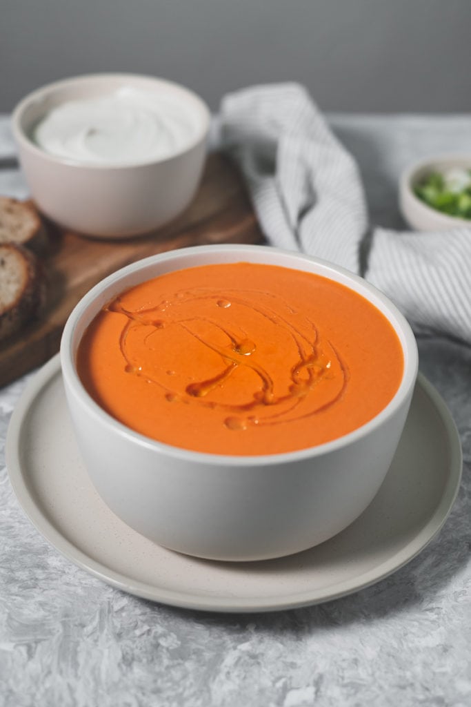 A full bowl of tomato soup with a fresh swirl of olive oil. Behind the soup is a linen cloth, a bowl of green onion, a bowl of cashew cream and a cutting board with slices of bread.