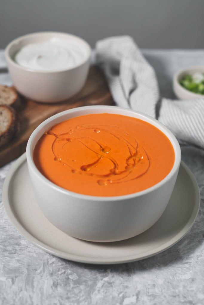 A full bowl of tomato soup with a fresh swirl of olive oil. Behind the soup is a linen cloth, a bowl of green onion, a bowl of cashew cream and a cutting board with slices of bread.