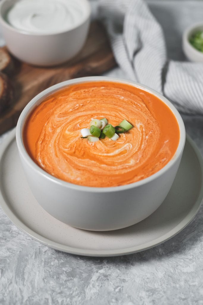 Homemade Tomato Soup swirled with cashew cream in a large stoneware bowl on a matching plate with a napkin, bowl of cashew cream, green onions and a board with slices of whole grain bread beside.