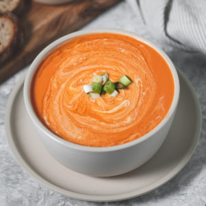 A large bowl of homemade tomato soup swirled with cashew cream and topped with green onion. Surrounding the bowl is a board with cashew cream, bread and on the other side is a linen napkin, spoon and bowl of chopped green onion.