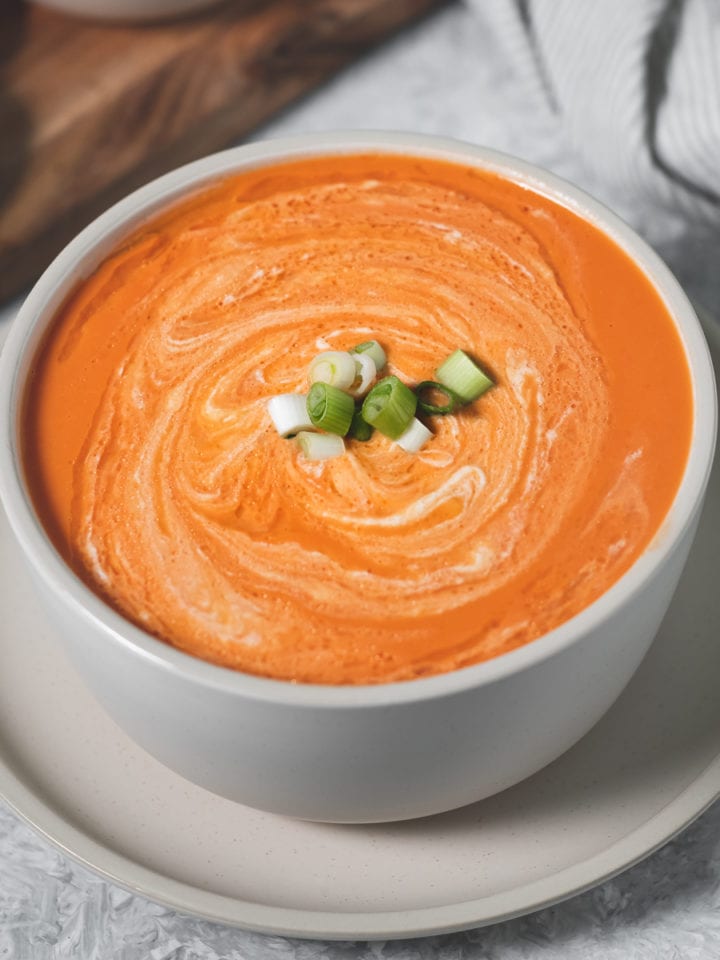 A large bowl of homemade tomato soup swirled with cashew cream and topped with green onion. Surrounding the bowl is a board with cashew cream, bread and on the other side is a linen napkin, spoon and bowl of chopped green onion.