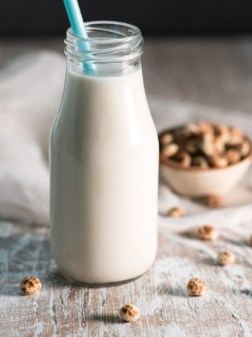 A single serve milk bottle full of tigernut milk with a blue straw sticking out and a bowl of tigernuts overflowing behind.