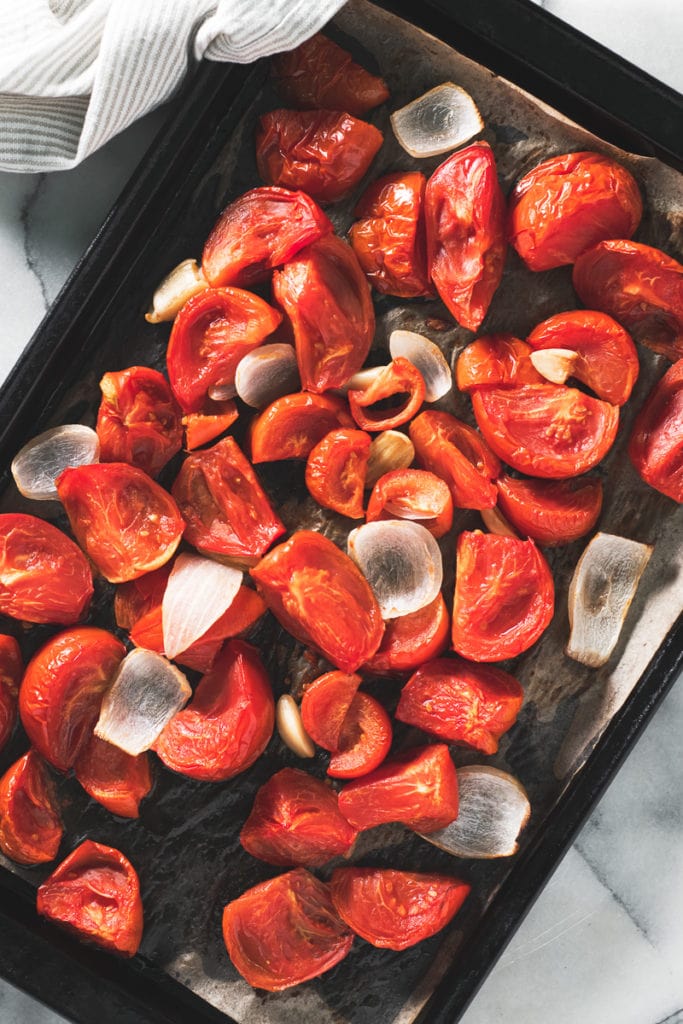 A sheet pan holding a spread of roasted tomato, onion and garlic with a dish cloth draped on the corner.