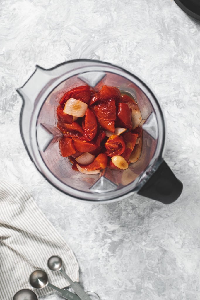 A blender jug filled with roasted tomatoes, onion and garlic.