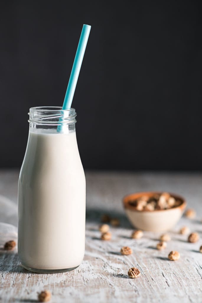 A jar of tigernut milk with a blue straw beside a spilled bowl of tigernuts.