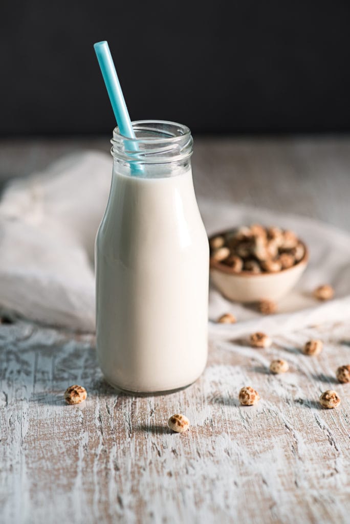 A bottle of tigernut milk with a blue straw in the middle of spilled tigernuts.