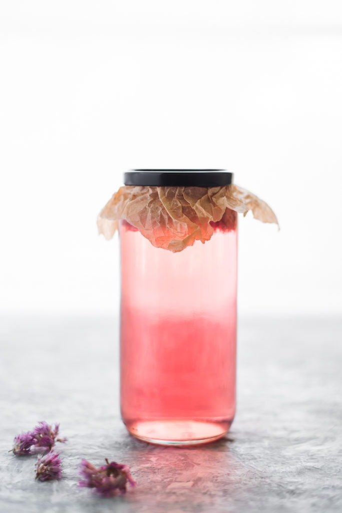 A jar of vibrant pink chive blossom vinegar with parchment paper sealed underneath the lid, and a few purple chive blossoms dropped in front.