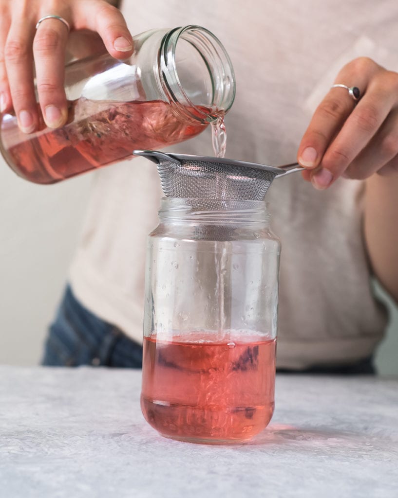 A person in a white t-shirt and jeans pouring infused chive blossom vinegar through a strainer into a clean jar.