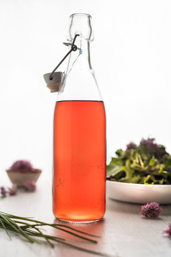 An open flip-top bottle filled with chive blossom vinegar, surrounded by a bunch of chives, a green salad and chive flowers.