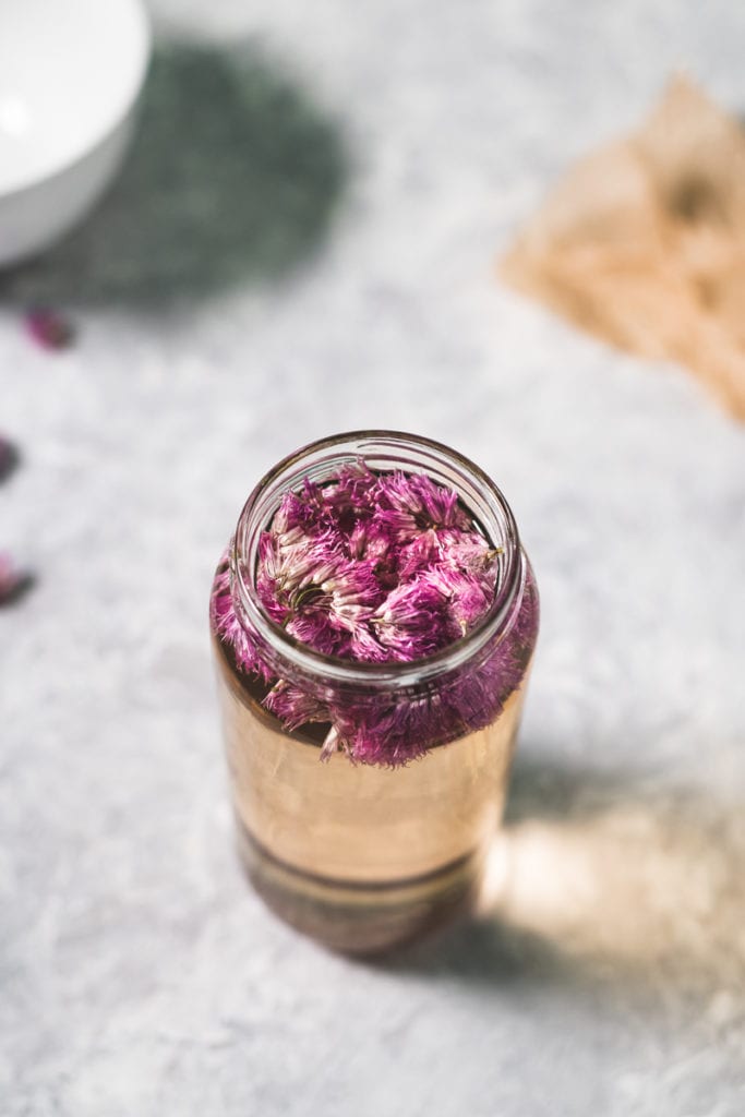 A jar filled with white wine vinegar and chive blossoms floating at the top, sitting in front of a bowl with chive blossoms spilling over and a piece of parchment paper.