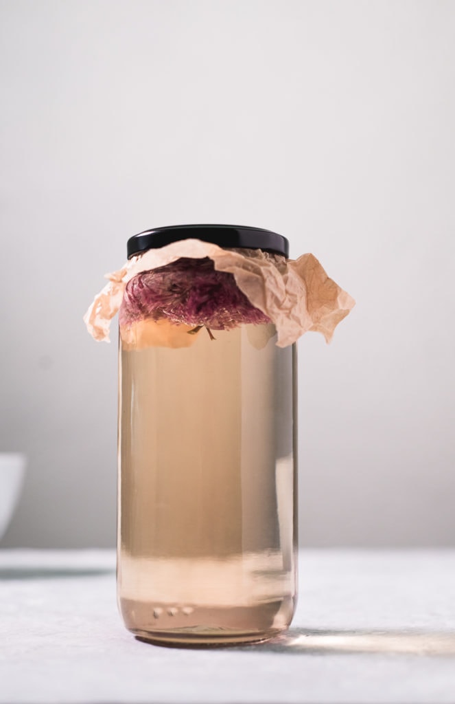 A jar filled with white wine vinegar and chive blossoms floating at the top, covered with a piece of parchment paper, sealed on underneath the lid.