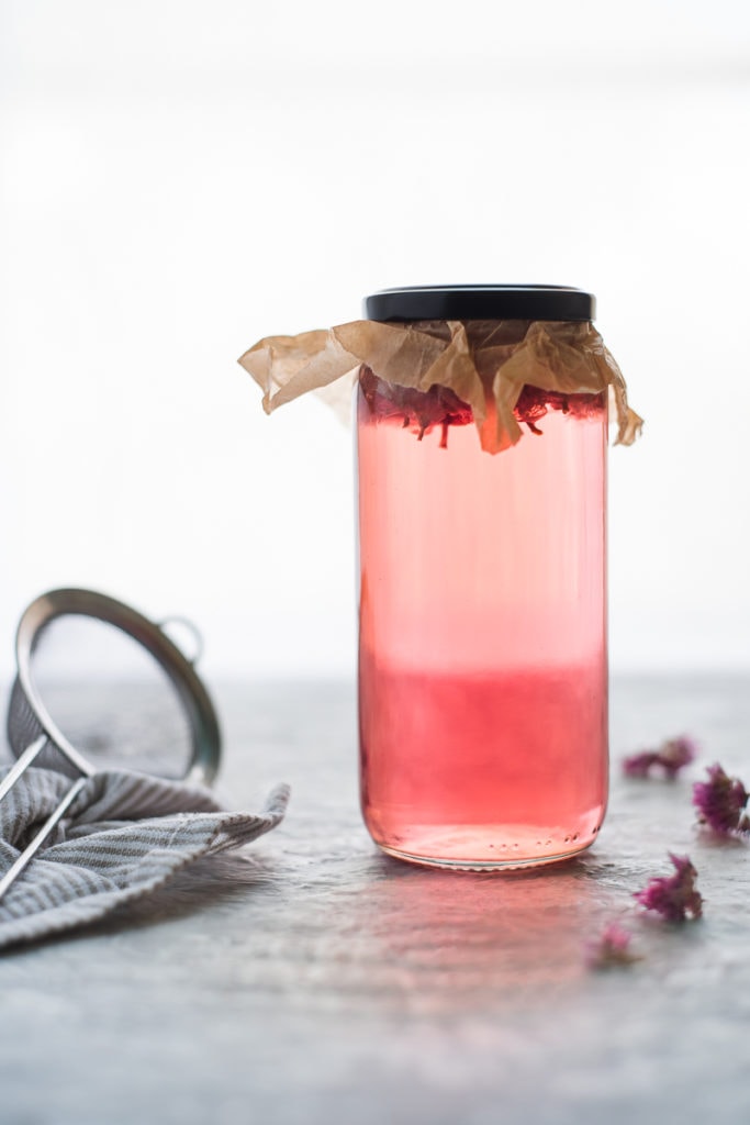 A jar of vibrant pink chive blossom vinegar with parchment paper sealed underneath the lid. On one side of the jar is a few dropped chive blossoms and on the other is a small strainer laying on top of a linen cloth.