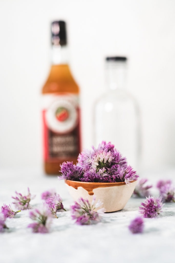 A small bowl overflowing with chive blossoms. Faded in the background is an empty glass bottle and a full bottle of apple cider vinegar.
