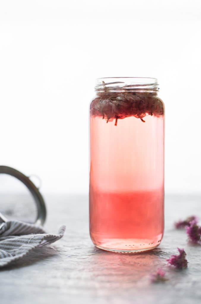 A gorgeous jar of pink chive blossom infused vinegar sitting in between dropped dried chive blossoms and a strainer resting on a striped linen cloth.