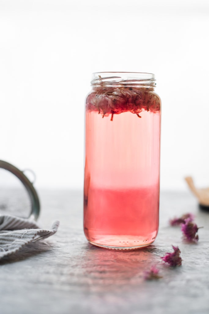 A jar of pink Chive Blossom Infused Vinegar with the chive flowers still floating atop. On on side of the jar is a small mesh strainer resting on a striped linen cloth. A few dropped dried chive flowers sit on the other side of the jar.