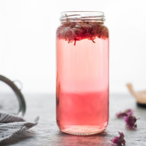 A jar of pink Chive Blossom Infused Vinegar with the chive flowers still floating atop. On on side of the jar is a small mesh strainer resting on a striped linen cloth. A few dropped dried chive flowers sit on the other side of the jar.