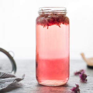 Chive blossoms floating at the top of a jar filled with pink vinegar, dried chive blossoms spread in to the right with a cloth and a sieve to the left.