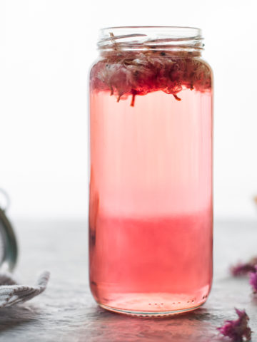 A jar of pink Chive Blossom Infused Vinegar with the chive flowers still floating atop. On on side of the jar is a small mesh strainer resting on a striped linen cloth. A few dropped dried chive flowers sit on the other side of the jar.