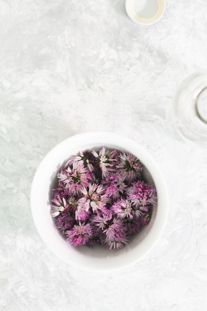 A white bowl filled with purple chive blossoms and an empty bottle sitting beside.