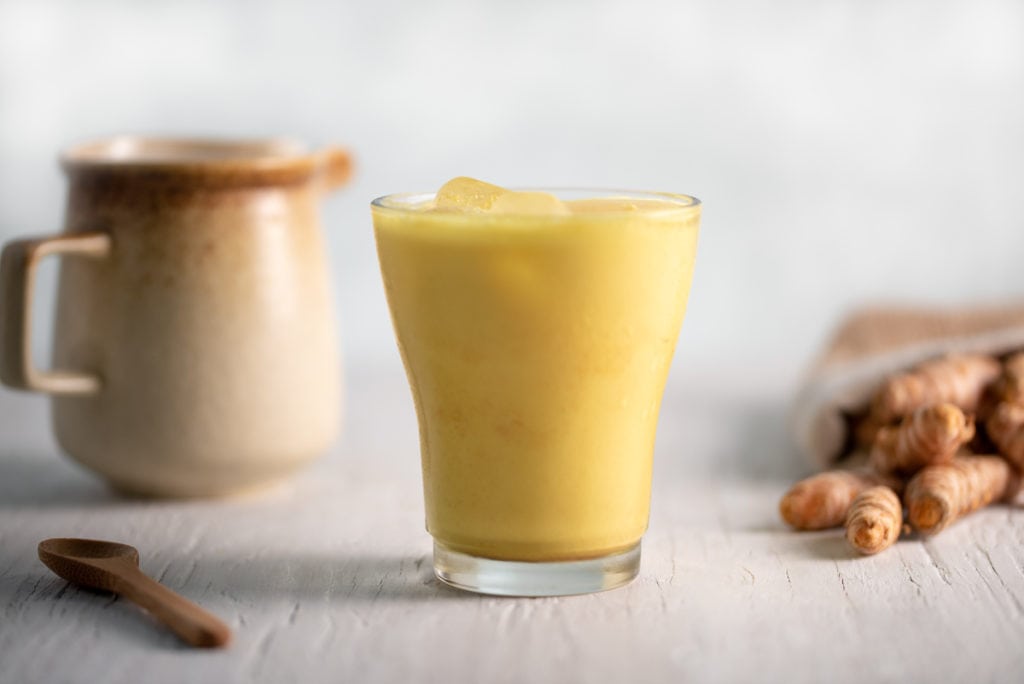 A frosty glass of iced golden milk centred between spilled turmeric and a ceramic milk jug.