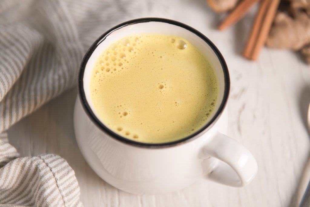 A mug of frothy golden milk in front of turmeric and cinnamon sticks.