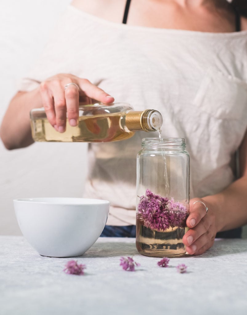 A person pouring white wine vinegar over chive blossoms in a jar, a white bowl sitting to the side and a few chive flowers sprinkled in front.