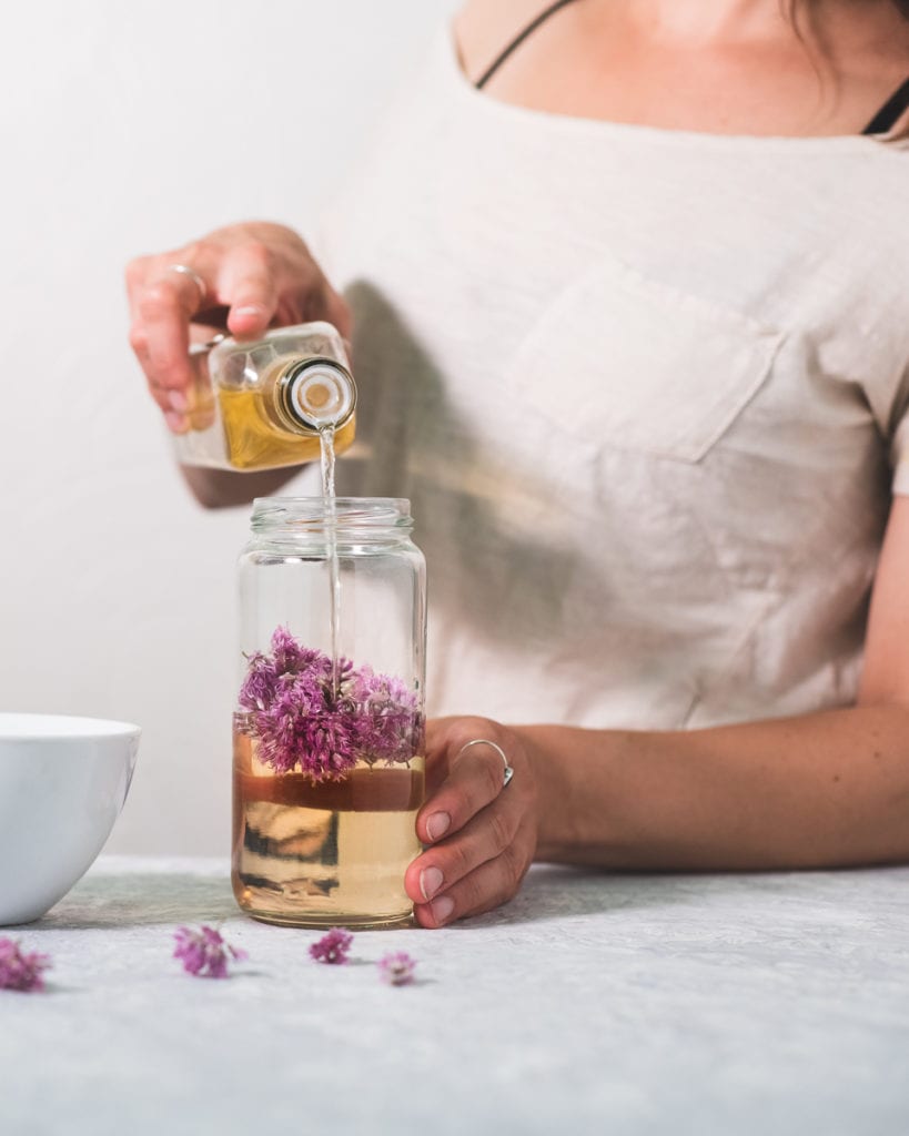 Person standing on an angle, pouring white wine vinegar into a jar with chive blossoms in it, a bowl to the side and a few chive flowers sprinkled about in front.