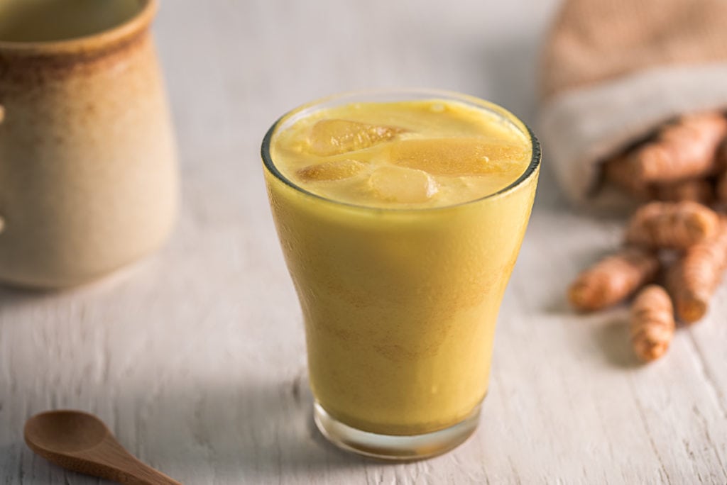 An iced golden milk latte in front of a ceramic milk jug and spilled turmeric root.