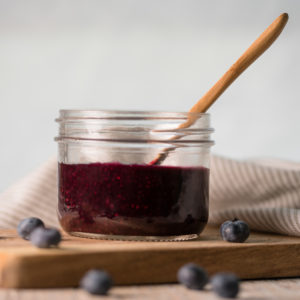 A jar of blueberry chia jam sitting on a cutting board with spilled blue berries all around.