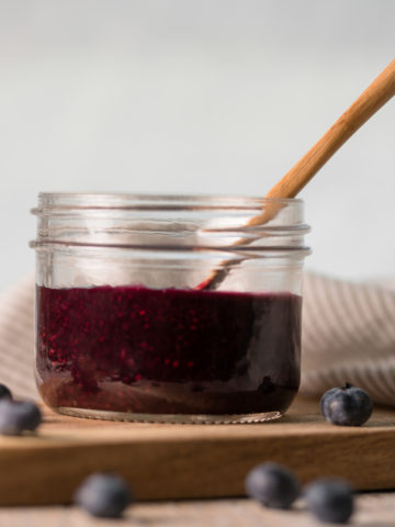 A mason jar of blueberry chia jam with a wooden spoon sticking out and blueberries sprinkled around.