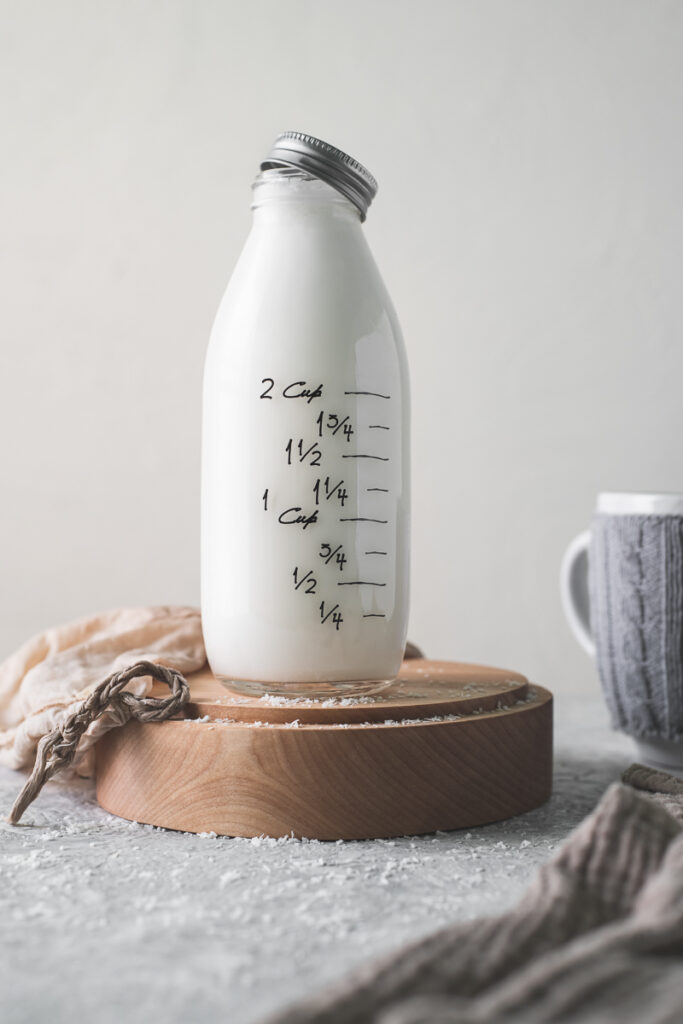 A full country milk bottle with the lid half-off, sitting on a wooden plate with a plant milk bag and shredded coconut spilt in front.