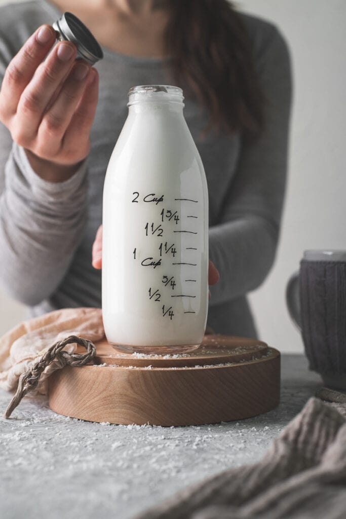 A woman lifting the cap off of a a very full country milk bottle with a freshly used plant milk bag resting beside and spilt coconut in front.