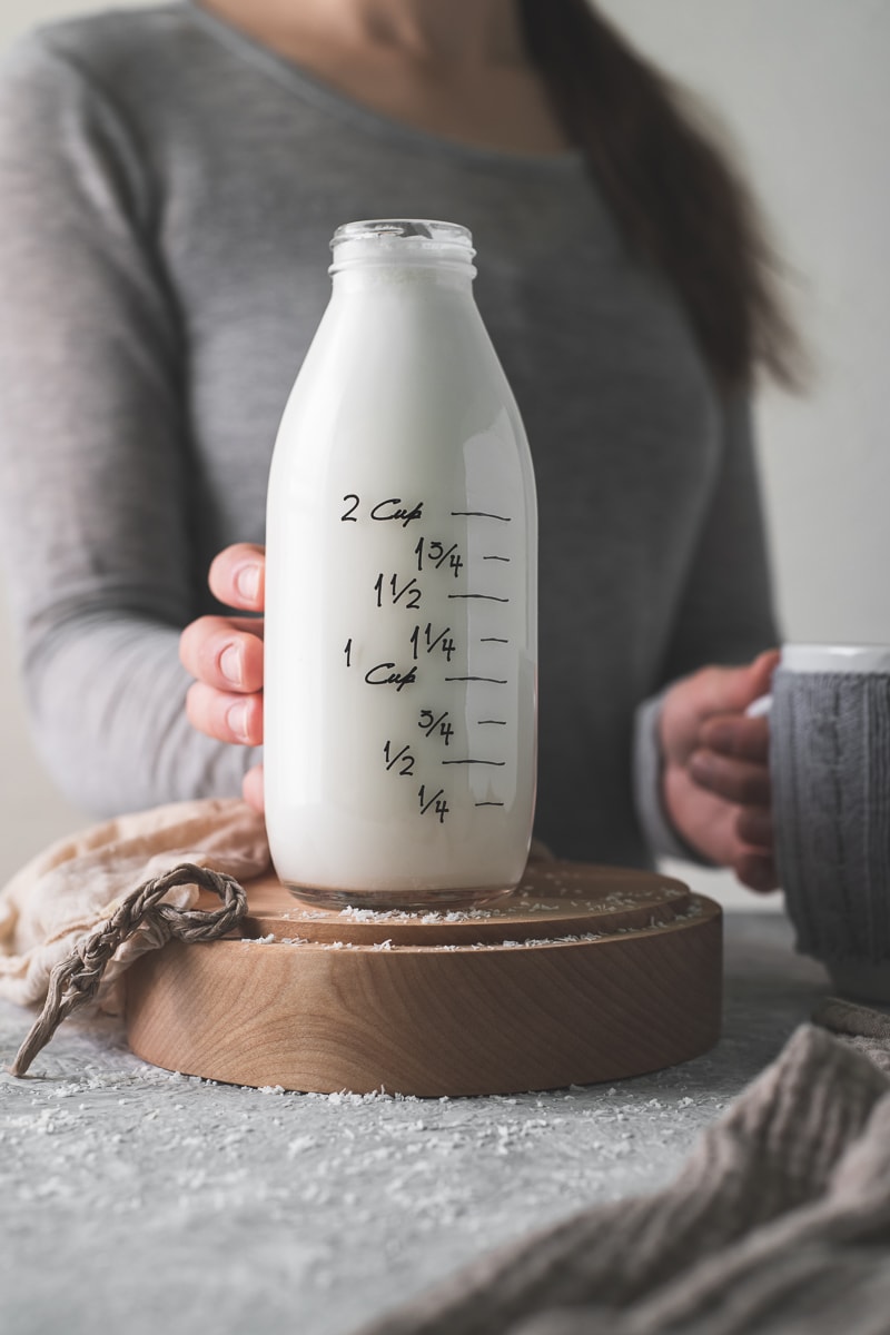 A woman holding a coffee mug reaching to pick up a full glass bottle of coconut milk.