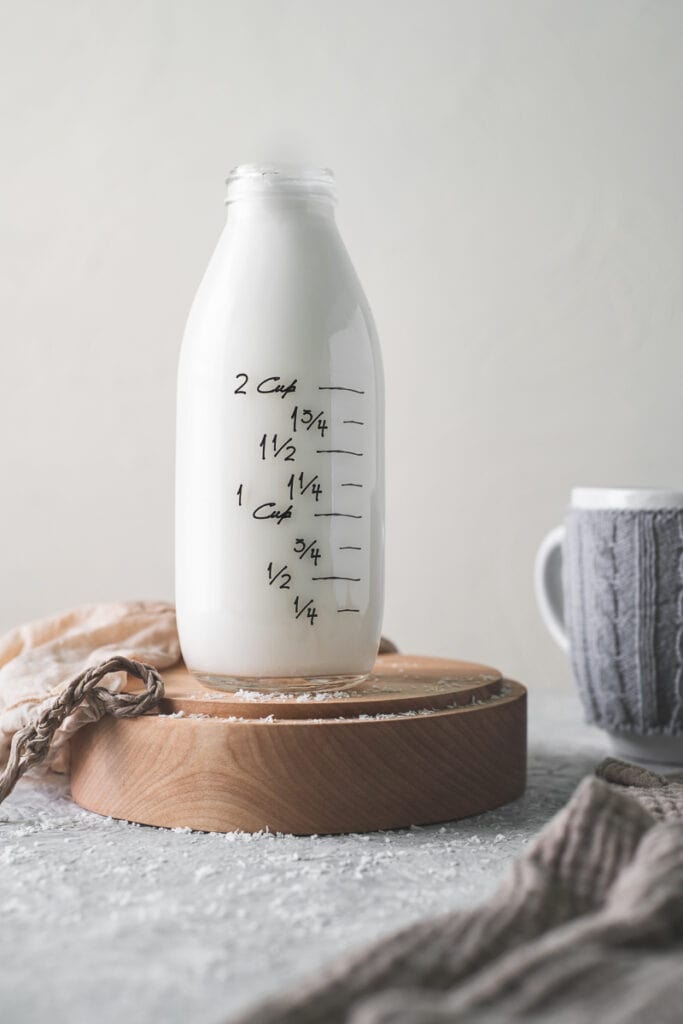 A small-mouthed country bottle filled to the top with freshly made coconut milk, sitting beside a coffee mug, linen cloth and a plant milk bag.