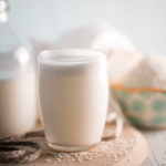 A glass of coconut milk sitting in front of a half full jar and an overflowing bowl of shredded coconut.