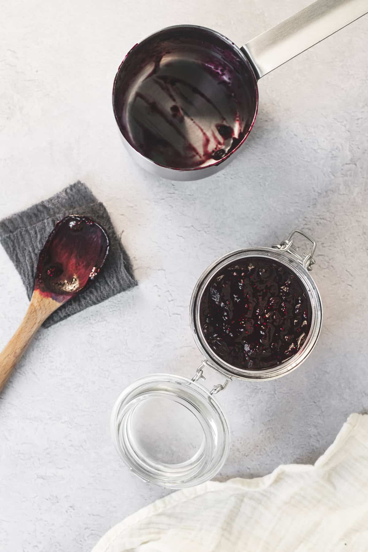 A dirtied wooden spoon and emptied sauce pan beside an open latch jar full of freshly poured blueberry chia jam.