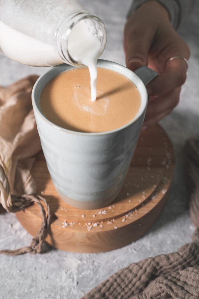 Coconut Milk being poured into a mug of tea with a cloth draped beside and shredded coconut spilt on the countertop.