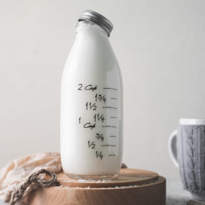 A full bottle of milk with the lid half-off, sitting on a wooden plate with a plant milk bag and shredded coconut spilt in front.