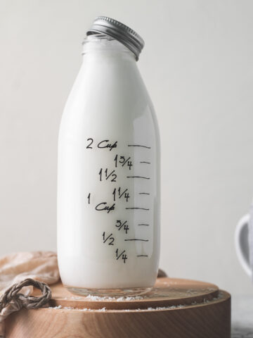 A full bottle of milk with the lid half-off, sitting on a wooden plate with a plant milk bag and shredded coconut spilt in front.
