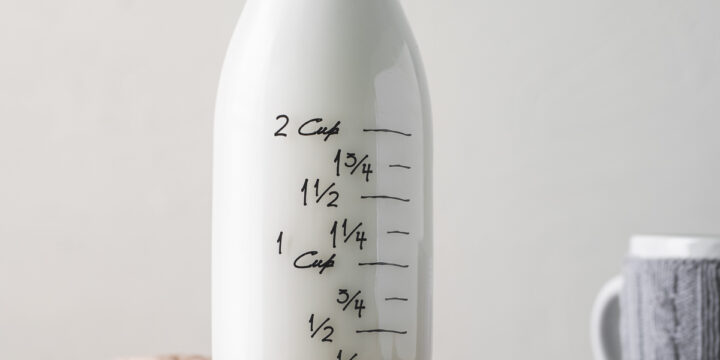 A full bottle of milk with the lid half-off, sitting on a wooden plate with a plant milk bag and shredded coconut spilt in front.