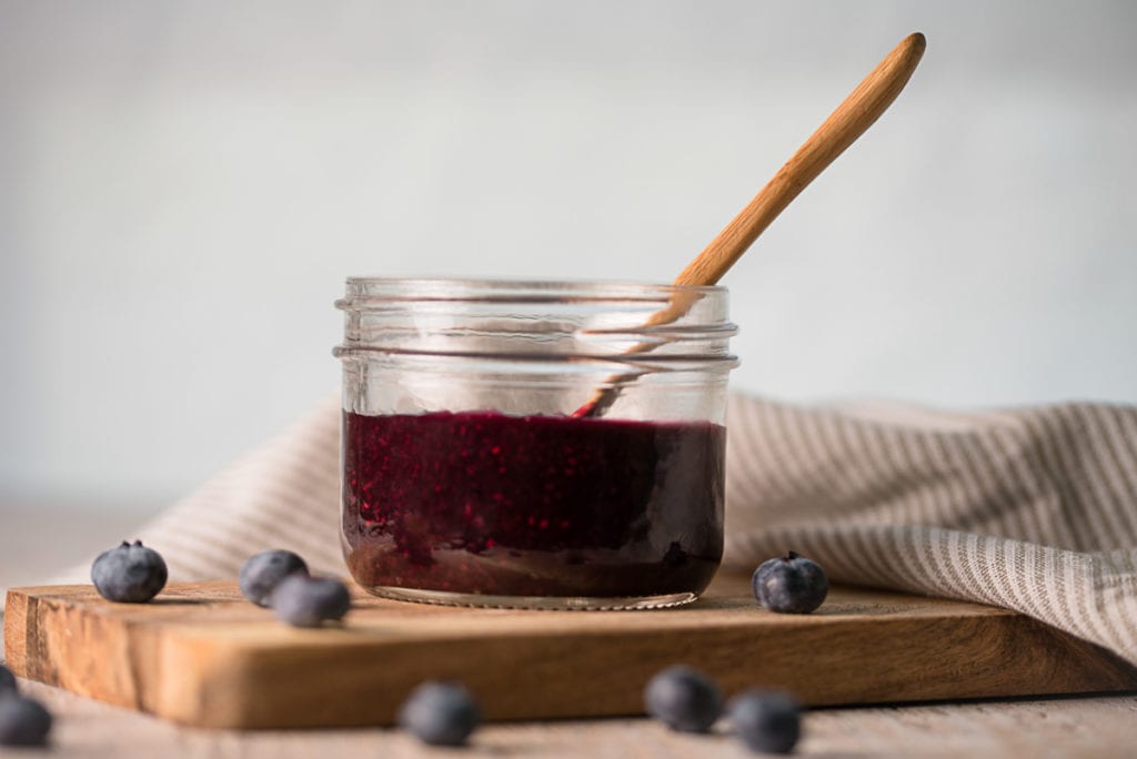 A jar of blueberry chia jam sitting on a cutting board with spilled blue berries all around.